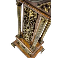 An Edwardian oak miniature longcase clock with a broken pediment and central brass finial, hood with applied brass spandrels supported on four reeded brass columns with Corinthian capitals, trunk with scrolled brass decoration on a corresponding stepped plinth raised on four ogee bracket feet, eight-day French timepiece movement with a later lever platform escapement, dial comprising a 3-1/4” enamel chapter ring with Arabic numerals and minute track, recessed applied decorative scroll work to the dial center, steel spade hands within a brass bezel and flat bevelled glass, rear wound and set.
With key.  
