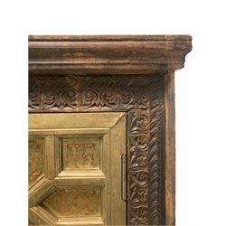 Indian hardwood cupboard, the front carved with a floral design, two doors with applied hammered and cut brass detail enclosing three shelves, raised on bun supports W117cm, H190cm, D62cm