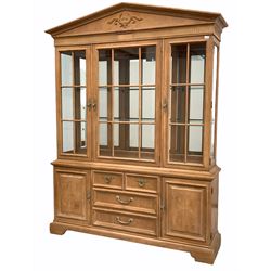 Large Georgian style light oak display cabinet/ bookcase, arched pediment centred by floral carved decoration, three glazed doors enclosing illuminated interior with fixed glass shelves over two cupboards and four drawers to centre, raised on shaped plinth base W160cm, H205cm