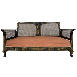 Early 20th century chinoiserie bergere settee, black lacquered with raised gilt decoration depicting figures and birds in landscape, caned back and sides, sprung seat upholstered in pink fabric with two cushions, on cabriole feet