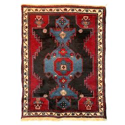 Persian Tuyserkan crimson ground rug, the central indigo geometric motif with stylised plant symbols, the spandrels as figure motifs, guarded ivory border with interconnected flower heads