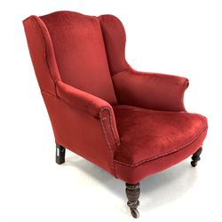 Edwardian wingback armchair, upholstered in red fabric, raised on turned front supports with castors 