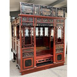 Late 19th/early 20th century Chinese hardwood opium or canopy bed, in red and black lacquered finish decorated with raised gilt and chinoiserie work, the pediment painted with flowers and figures, decorated all-over with carved panelled depicting various figural scenes and flowers, the windows decorated with fretwork surrounds and carved flower heads, fitted with three drawers and wooden latted base