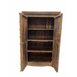 Indian hardwood cupboard, the front carved with a floral design, two doors with applied hammered and cut brass detail enclosing three shelves, raised on bun supports W117cm, H190cm, D62cm