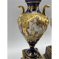 Pair of 'Vienna' porcelain urns, of typical urn form with gilt scroll handles, cobalt blue ground and gilt bands painted with continuous scenes of 'Paris and Helena' & 'Diana im Bade', the other painted with similar scenes, titled beneath, raised on square bases, beehive mark in blue, H23cm