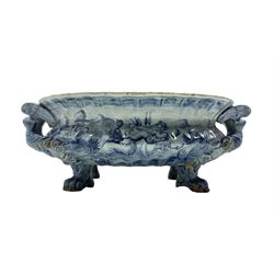 17th/ 18th century Italian Maiolica blue and white cistern, possibly Savona, of oval gadrooned form with two handles formed as angels branching into corkscrew coils, the bowl painted with Putti and figures in a landscape, supported by four paw feet, L55cm, W36cm, H19cm