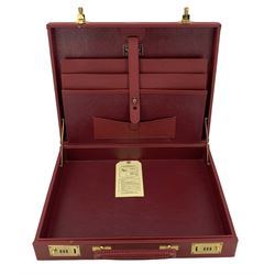 Late 20th century Aston Martin red leather suitcase and an attaché, by Tanner & Krolle, with brass combination locks and mounts, stamped with gilt Aston Martin emblem, the suitcase with red silk lined interior with two pockets, the attaché fitted with a folio, suitcase 59cm wide, attaché 42.5cm wide (2)