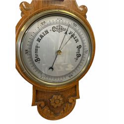 An English 1930’s solid oak carved hall barometer in a scroll shaped carved case with applied floral and leaf carving, compensated Aneroid movement, eight-inch silver painted dial measuring barometric air pressure from twenty-six to thirty-one point nine inches, weather predictions written in black upper and lower case gothic script with a blue steel indicating hand and brass recording hand, brass dial bezel with flat bevelled glass, mercury thermometer enclosed in a glazed rectangular box recording temperature in degrees Fahrenheit and Celsius on a white porcelain register, dial inscribed “Made in England”