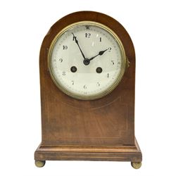 An Edwardian mantle clock in an arched mahogany case inlaid with satinwood stringing on a moulded plinth with ball feet (one missing), arched rear door with brass sound fret, eight-day French striking movement with a recoil anchor escapement striking the hours and half hours on a coiled gong, with a white convex enamel dial, upright Arabic numerals and minute track, steel spade hands within a cast bezel and convex glass. With pendulum.
H 26 W 19 D14