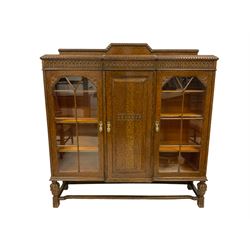 Early 20th century oak bookcase cabinet, breakfront with raised back, frieze carved with arcade decoration, fitted with two flanking astragal glazed doors and central panelled cupboard, brass handles in the shape of pharaoh masks, raised on turned supports with gadroo design, united by shaped stretcher