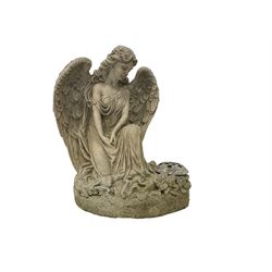 Composite stone garden ornament or memorial statue in the form of a Classically draped winged angel kneeling, on a naturalist base with flowers