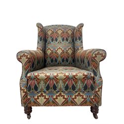 Late 19th century mahogany framed wingback armchair, scrolled arms and sprung back and seat, upholstered in Art Nouveau design Liberty 'Ianthe' fabric, raised on turned front supports terminating in brass and ceramic castors