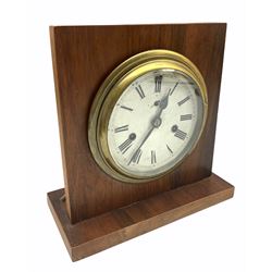 20th century circular brass cased clock with a twin train spring driven movement in the style of a bulkhead clock, 17.78 cm diameter brass case with a 15.24 cm diameter brass bezel, 12.7 cm diameter white paper dial with Roman numerals and minute track, spade hands and second hand, housed in a mahogany display stand, with key.