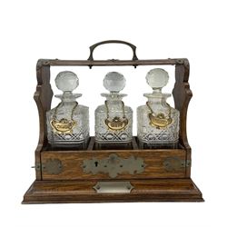 Early 20th century oak three bottle tantalus with originally plated mounts and three cut glass square section decanters, H35cm x W37cm 