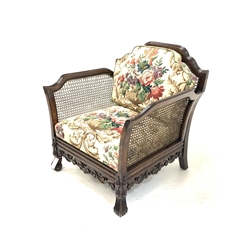 Early 20th century polished beech carved bergere armchair, shaped cresting rail and out swept arms over scrolling moulded decoration to seat rails, on scrolling knop supports, upholstered in vintage Sanderson linen