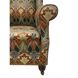 Late 19th century mahogany framed wingback armchair, scrolled arms and sprung back and seat, upholstered in Art Nouveau design Liberty 'Ianthe' fabric, raised on turned front supports terminating in brass and ceramic castors