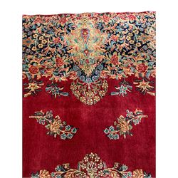 Persian Kerman crimson ground carpet, the field with a large central floral medallion, the flower motifs with a pale indigo outline, surrounded by bouquets within the plain field,  the border decorated with closely woven flower heads connected by scrolling acanthus leaves
