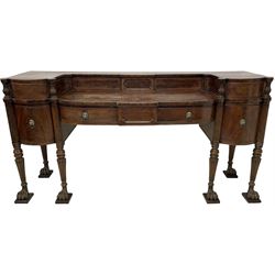 An impressive Irish George III mahogany sideboard, raised back fitted with sliding compartments and two drawers with reed carved balusters and applied cock-beading, fitted with three shallow central drawers and two flanking deep drawers, the central drawer with shaped figured panel, reeded uprights and turned and reeded tapering supports with carved paw feet on moulded blocks, brass lion masks with foliate garland ring handles