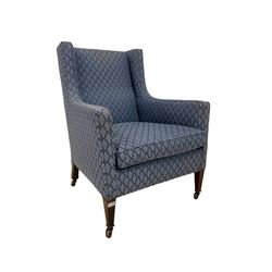 Edwardian wingback armchair, sprung back and seat upholstered in foliate patterned blue fabric, raised on mahogany tapered supports terminating in ceramic castors 