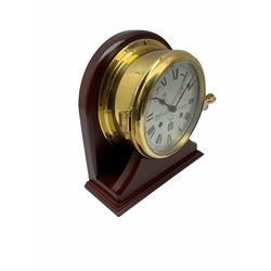 Eight-day 20th century brass cased ships bulkhead clock mounted on a mahogany effect display stand, with roman numerals and minute track, fast slow regulation, bells on/off control, black spade hands with lockable bezel, twin train spring driven balance wheel movement sounding the ships watches on a bell.