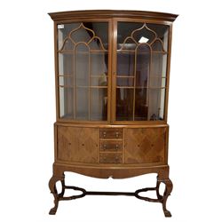 Early 20th century inlaid walnut bow-front display cabinet, dentil frieze over two astragal glazed doors enclosing two shelves, surrounded by chequered stringing, the base fitted with three central crossbanded drawers, flanked by two marquetry cupboards, shaped apron over cabriole supports, united by moulded shaped X-stretcher