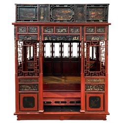 Late 19th/early 20th century Chinese hardwood opium or canopy bed, in red and black lacquered finish decorated with raised gilt and chinoiserie work, the pediment painted with flowers and figures, decorated all-over with carved panelled depicting various figural scenes and flowers, the windows decorated with fretwork surrounds and carved flower heads, fitted with three drawers and wooden latted base