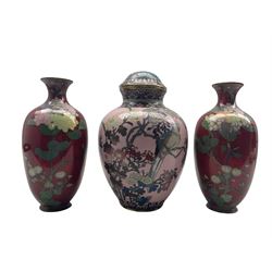 Japanese Cloisonne vase and cover decorated with an exotic bird perched on a branch amongst wisteria, iris and chrysanthemums on a pale pink ground, with associated cover, H29cm, together with a pair of Japanese Cloisonne vases on red ground (3)