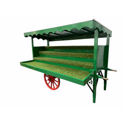 Florists market hand drawn barrow in green and red livery, the sloped top with shaped apron over four simulated grass covered staggered tiers and storage well, cast iron leaf suspension and iron bound twelve spoke wheels, L290cm