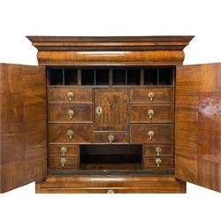 George I walnut cabinet on chest, the projecting stepped and moulded cornice over single cushion drawer, the two feather banded cabinet doors opening to reveal a fitted interior comprising of four pigeonholes concealing secret drawers, eleven assorted drawers with a central cupboard, the figured walnut facias with feather banding and drop pull handles in the form of masks, the lower section fitted with three graduating drawers with moulded facias and press brass plates and escutcheons, raised on shaped bracket feet