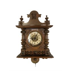 German - Edwardian 8-day striking wall clock in an oak case, with a shaped pediment, turned columns and finials, two part dial with a gilt centre, gothic hands and Arabic numerals, two-train movement striking the hours and half-hours on a coiled gong, visible gridiron pendulum with a visible brass bob.