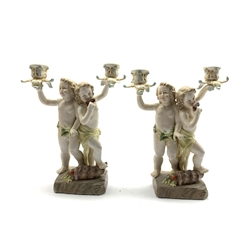  Two 20th century Continental porcelain two branch candelabra modelled as two cherubs playing pan pipes with a cornucopia overflowing with fruit on the ground,  H29cm   