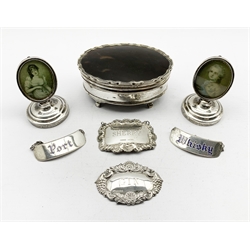 Silver oval dressing table box with hinged tortoiseshell cover on shaped supports W10cm Birmingham 1920 Maker Mappin and Webb, pair of silver menu holders inset with a portrait print Chester 1904, pair of silver and blue enamel decanter labels and two others