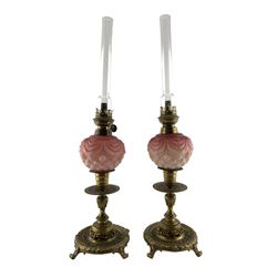 Pair of late Victorian ornate brass candlesticks by William Tonks, later converted to oil lamps, with pink satin glass reservoirs, clear glass funnels and brass burners, on four scroll supports, candlesticks impressed W.T & S, H52cm overall