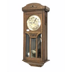 A 1930’s oak cased wall clock with an eight-day movement striking the hours and half-hours on a coiled gong, with a silvered dial and upright Arabic numerals, steel spade hands and brass spun bezel, the case with shaped pediment and applied carving, full length door with three bevelled glass panels and visible spun brass pendulum. With key.