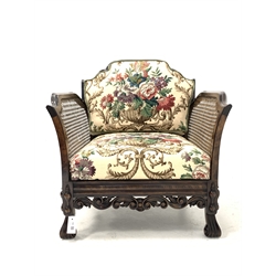 Early 20th century polished beech carved bergere armchair, shaped cresting rail and out swept arms over scrolling moulded decoration to seat rails, on scrolling knop supports, upholstered in vintage Sanderson linen