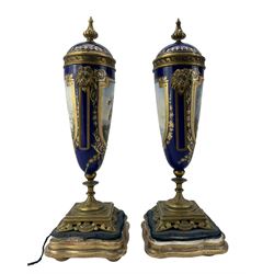Pair of 19th century Sevres style porcelain and gilt metal urns and covers, the cobalt blue ground painted with  two oval reserves of a courting couple and landscapes, each within jewelled and gilt borders, twin lion mask handles and square bases with pierced supports, on giltwood and velvet serpentine bases with bun feet, H28cm 